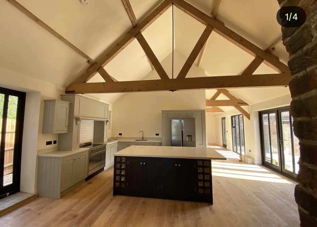 A grey painted barn kitchen with a vaulted ceiling, exposed timber trusses and a dark island, freshly decorated