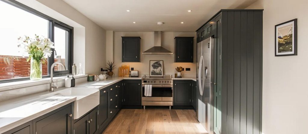 A dark green shaker kitchen with a white worktop and a butler sink, the walls and ceiling painted off white