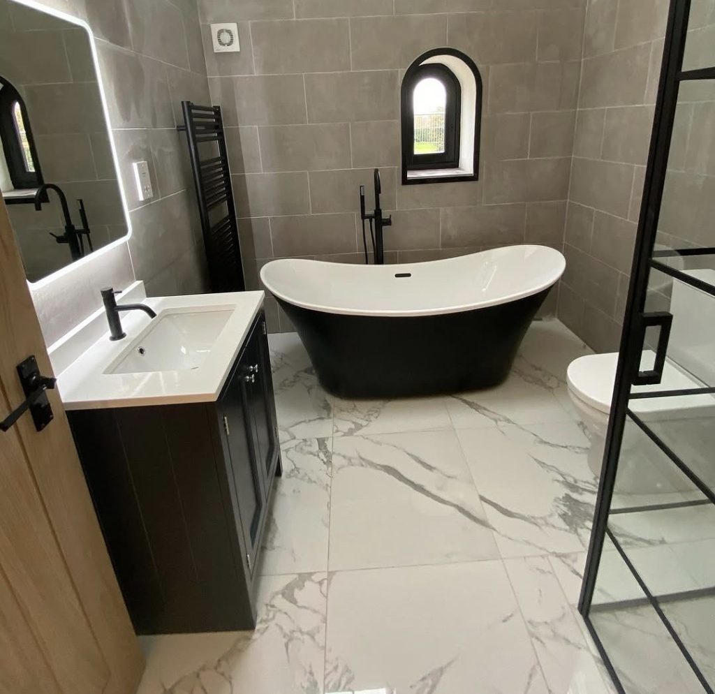 A bathroom tiled in large grey porcelain with a marble effect floor, a black freestanding bath and an arched window