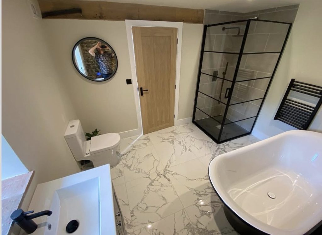 An en suite with a marble effect tiled floor, a black framed corner shower and a freestanding bath beneath an exposed beam