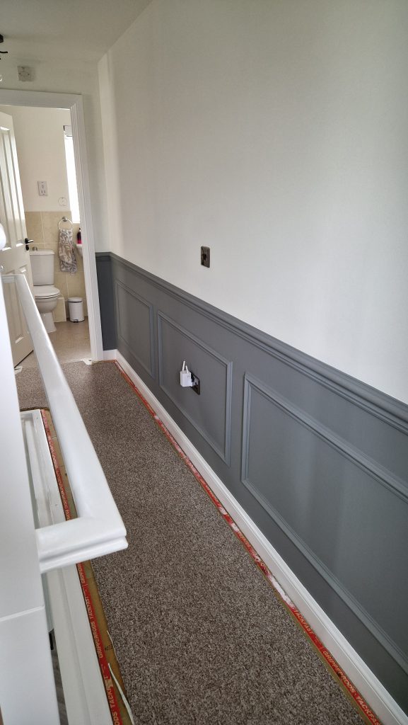 A landing with grey panelled wainscoting below pale painted walls, and freshly painted white skirting and architrave