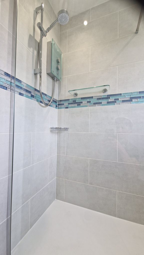 A shower tiled in large pale grey tiles, with a band of blue and green glass mosaic running round the room