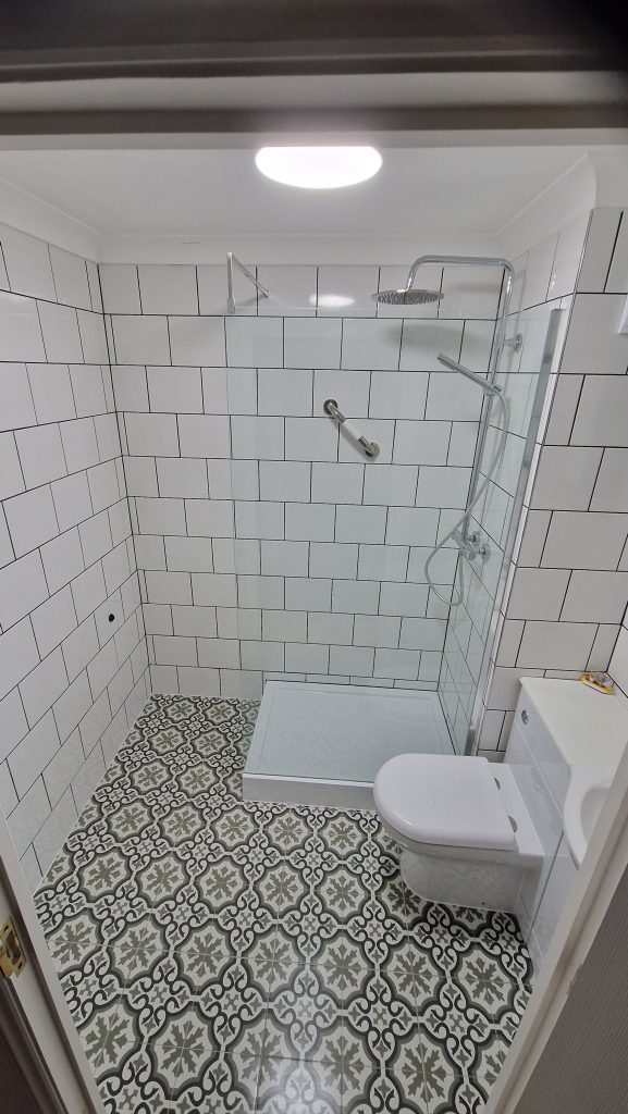 A shower room tiled in white square tiles with dark grout, above a grey and white patterned floor